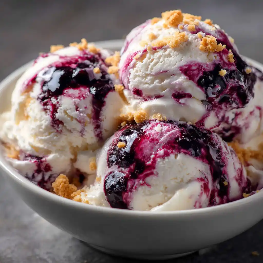 three scoops of blueberry cheesecake ice cream in a bowl with blueberry swirl and graham cracker crumbs